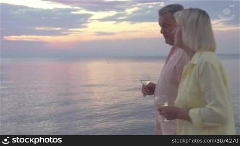 Steadicam shot of mature couple walking by the sea at sunset with wine glasses in hands.