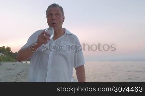 Steadicam shot of mature man walking along the sea, writing something in smartphone and drinking from the wine glass.