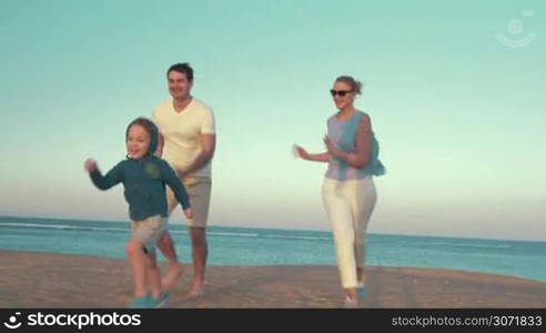 Steadicam shot of mother and fathrer running a race with son on the beach. Boy running up to broken buoy and kicking it, parents applauding to little winner