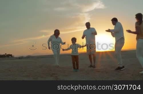 Steadicam shot of parents, little child and grandparents spending evening on the beach. They dancing on the sand against bright sunset. Happy family vacation