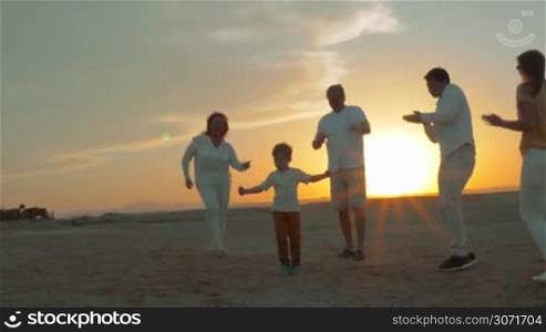 Steadicam shot of parents, little child and grandparents spending evening on the beach. They dancing on the sand against bright sunset. Happy family vacation