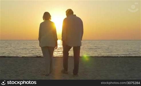 Steadicam shot of senior couple coming to the beach to see the beauty of sunset. They embracing and looking at sea and sundown
