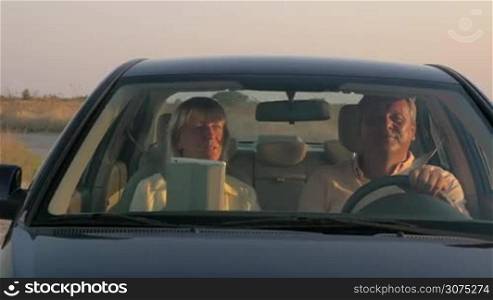 Steadicam shot of senior couple having a slow car ride in rough country at sunset. Man driving and woman using tablet computer