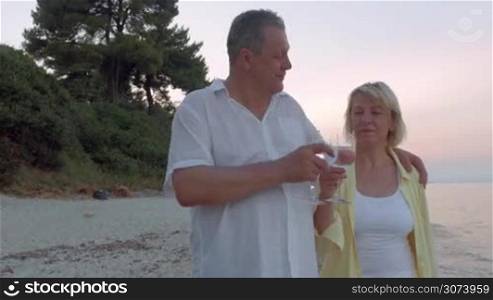 Steadicam shot of senior couple having outdoor walk by the sea and drinking water. Man embracing woman and they enjoying quiet evening at the seaside