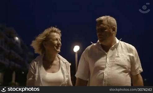 Steadicam shot of senior couple having walk at windy night on resort and taking selfie on mobile