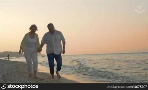 Steadicam shot of senior couple holding hands and running along the sea coast at sunset