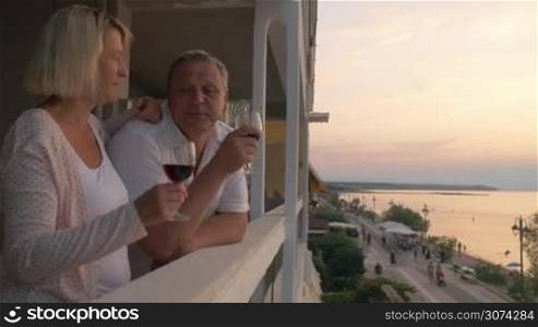 Steadicam shot of senior family couple spending evening on drinking wine on hotel balcony on resort. They looking at scenic seascape with sunset