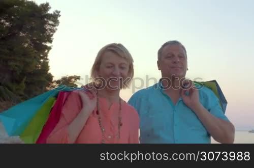 Steadicam shot of senior man and woman having relaxing evening walk on the coast, they carrying bags on the shoulders. Shopping on vacation