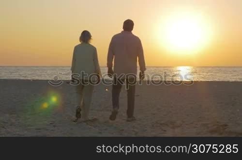 Steadicam shot of senior man and woman holding hands and walking to the water. They enjoying sunset over sea in hugs
