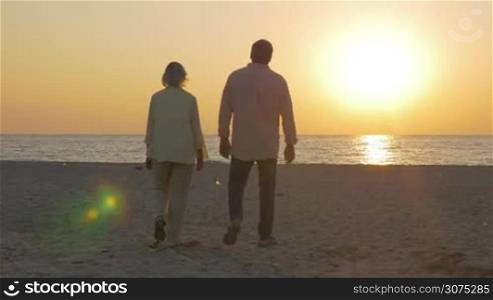 Steadicam shot of senior man and woman holding hands and walking to the water. They enjoying sunset over sea in hugs