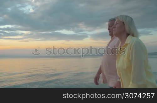 Steadicam shot of senior man and woman walking along the quiet sea at sunset. Loving couple holding hands and embracing