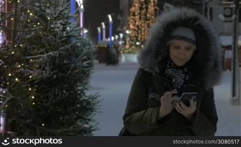 Steadicam shot of smiling woman typing on mobile as she walking in the street with Christmas illumination. Winter scene with holiday atmosphere
