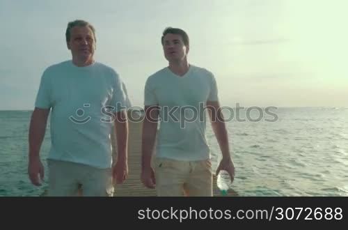 Steadicam shot of two friends or father and adult son having conversation while walking along the wooden pier in the sea