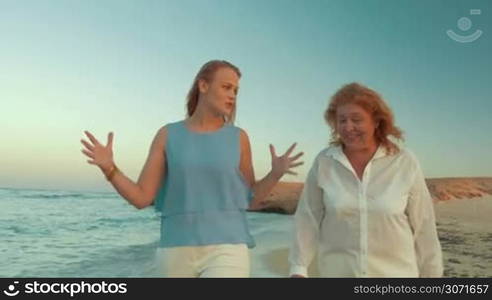 Steadicam shot of two women, young and mature, or mother and daughter walking alongside the sea and talking to each other.