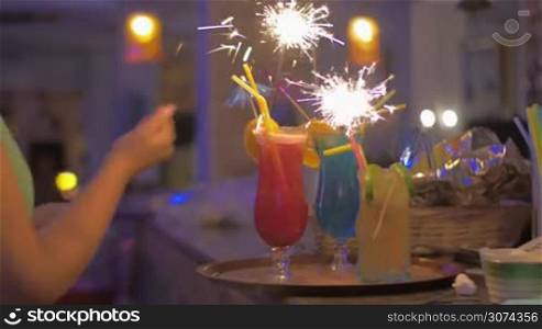 Steadicam shot of waitress lighting sparklers in glasses with cocktails and serving them on the tray.