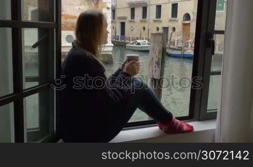 Steadicam shot of woman sitting alone on the windowsill. Window is open and she looking at Venice scene while having hot tea