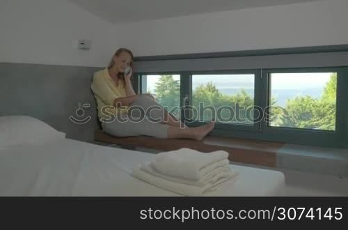 Steadicam shot of young blond woman having a phone talk while sitting by the window in hotel room. Beautiful nature view from the window