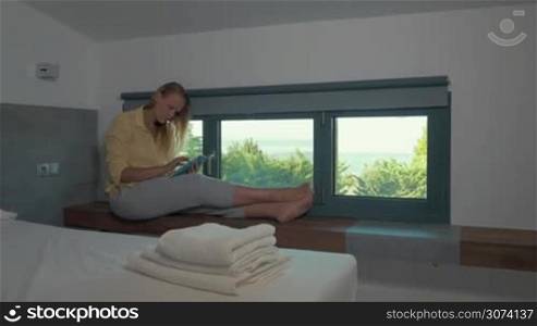 Steadicam shot of young blond woman sitting on windowsill in hotel room and using tablet computer. Green nature scene in the window