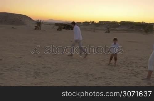 Steadicam shot of young family kicking a little ball on a beach at twilight.