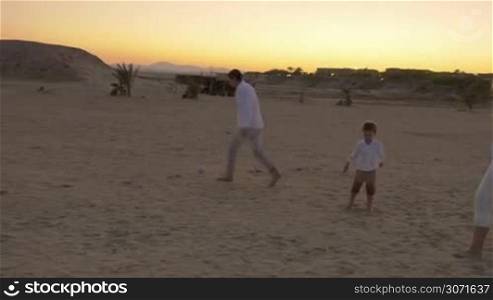Steadicam shot of young family kicking a little ball on a beach at twilight.