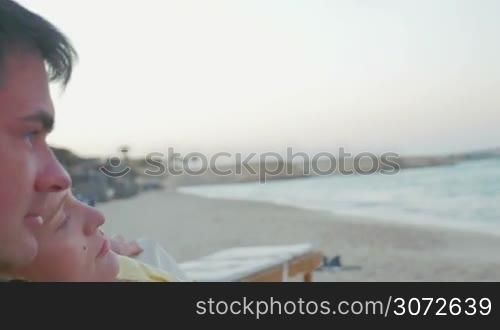 Steadicam shot of young family lying on the chaise longue on the shore. Relaxed parents enjoying seascape while their little son playing with smart phone