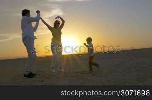 Steadicam shot of young happy parents and little son having fun while dancing on the beach at sunset. Bright evening sun shining in background. Summer vacation with family