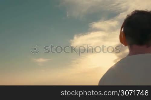 Steadicam shot of young man and woman having enjoyable evening outdoor. They listening to music in wireless headphones and admiring scenic view of sea and sky