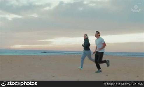 Steadicam shot of young man and woman having evening training on the beach. They jogging and listening to music in wireless headphones