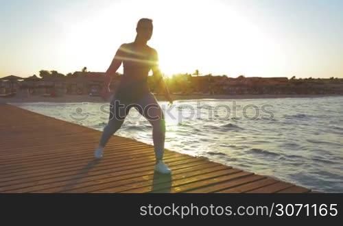 Steadicam shot of young woman doing lunge during her morning exercises outdoors. She is lighted by rising sun.