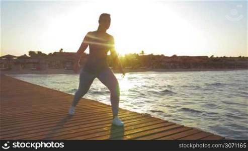 Steadicam shot of young woman doing lunge during her morning exercises outdoors. She is lighted by rising sun.