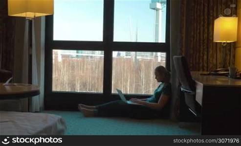 Steadicam shot of young woman using laptop in hotel. She sitting on the floor by the window in cosy room, chatting on computer and enjoying city view
