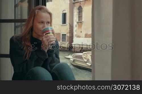 Steadicam shot of young woman with wistful look drinking hot coffee sitting on the windowsill at home by Venice canal. She enjoying outside view