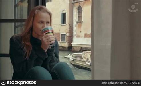 Steadicam shot of young woman with wistful look drinking hot coffee sitting on the windowsill at home by Venice canal. She enjoying outside view