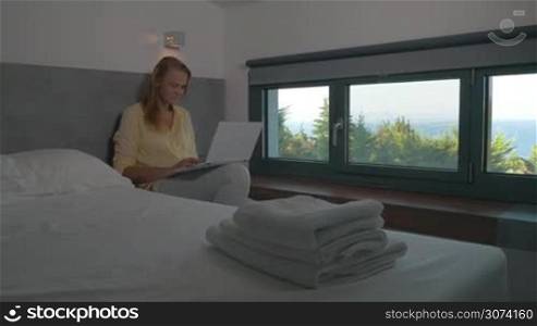Steadicam shot of young woman working with laptop in hotel room and receiving a video call. She having excited talk and showing nature view from the window