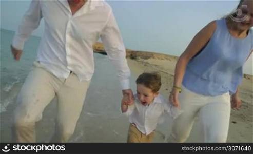 Steadicam slow motion of a young family running along the sea and holding hands. Little boy is jumping and hanging at poise with parents&acute; arms.