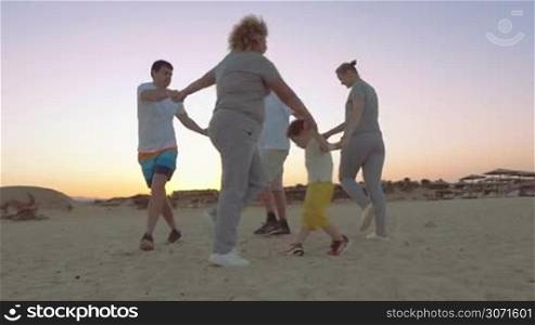 Steadicam slow motion shot of a big family holding hands and dancing round dance on the evening beach.