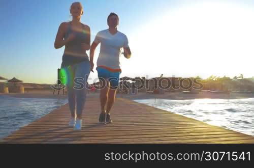 Steadicam slow motion shot of a young couple jogging along the wooden pier. Rising sun is lighting their backs.