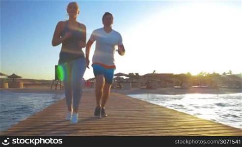 Steadicam slow motion shot of a young couple jogging along the wooden pier. Rising sun is lighting their backs.