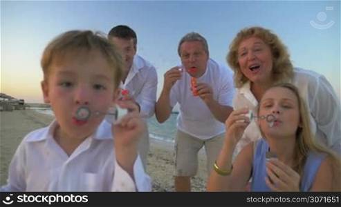 Steadicam slow motion shot of people blowing soap bubbles. Little boy is doing it on the foreground and his parents and grandparents are on the background.