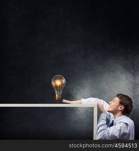 Steal the idea. Young man looking from under table at glass light bulb