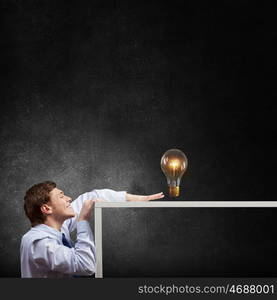 Steal the idea. Young man looking from under table at glass light bulb