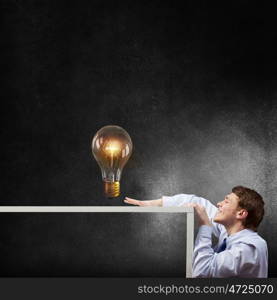 Steal the idea. Young man looking from under table at glass light bulb