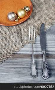 Steel fork and knife and Christmas balls on a ceramic plate are located on the rough wooden table, covered with coarse matting