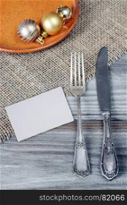 Steel fork and knife and Christmas balls on a ceramic plate are located on the rough wooden table, covered with coarse matting, with white paper card