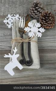 Steel fork and knife handmade lie on a linen napkin tied with rough twine as well as two pine cones and a white felt toy in the form of snowflake and deer are located on the rough wooden table