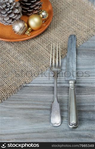 Steel fork and knife, two pine cones and Christmas balls on a ceramic plate are located on the rough wooden table, covered with coarse matting