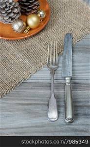 Steel fork and knife, two pine cones and Christmas balls on a ceramic plate are located on the rough wooden table, covered with coarse matting