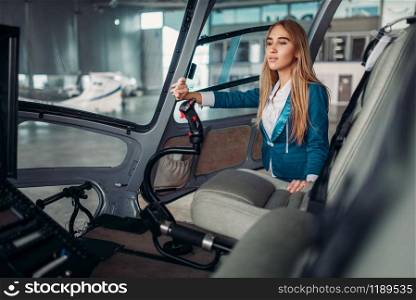Stewardess in suit poses in helicopter in hangar, view from cabin. Air hostess in uniform near copter. Private airline. Stewardess poses in helicopter, view from cabin