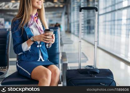 Stewardess with coffee and suitcase sitting on seat in waiting area in airport. Air hostess with baggage, flight attendant with hand luggage, aviatransportations job. Stewardess with coffee on seat in waiting area