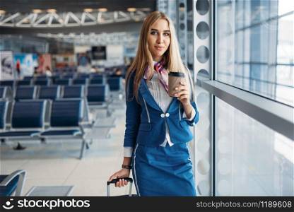 Stewardess with suitcase and coffee against window in airport. Air hostess with baggage in waiting room, flight attendant with hand luggage, aviatransportations job. Stewardess with hand luggage and coffee in airport